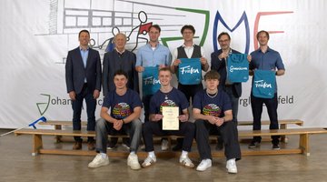 Drei Schüler auf einer Bank, dahinter für fünf Personen, die zum Gewinn des Schülerwettbewerbs gratulieren.